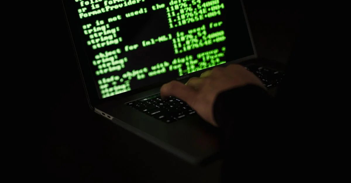 Silhouette d'un hacker tapant sur un ordinateur portable dans le noir avec un code vert à l'écran.